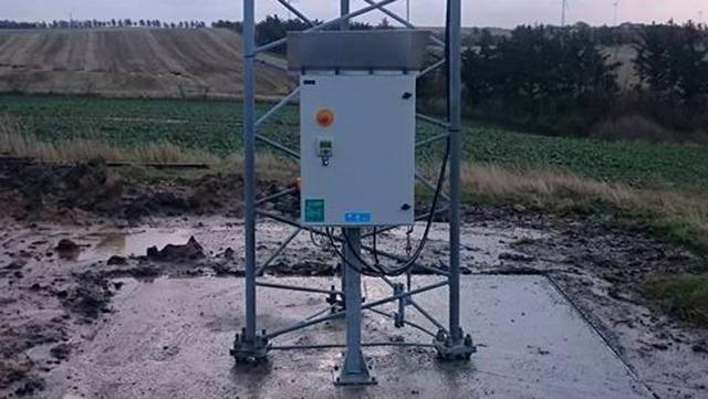 En målerstation på en metalstolpe i et mudret landskab med marker i baggrunden.