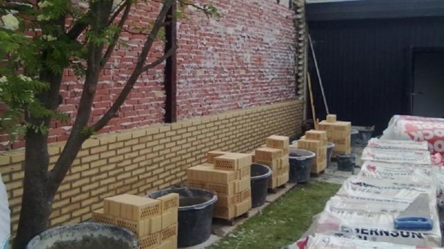 Mur med rød mursten og en række kasser og potter langs jorden, omgivet af byggematerialer.