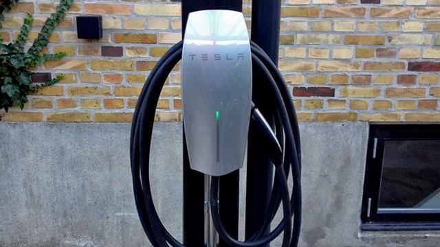 Ladepunkt til elbil fra Tesla med en sort ladekabel hængende ved en mur af mursten.