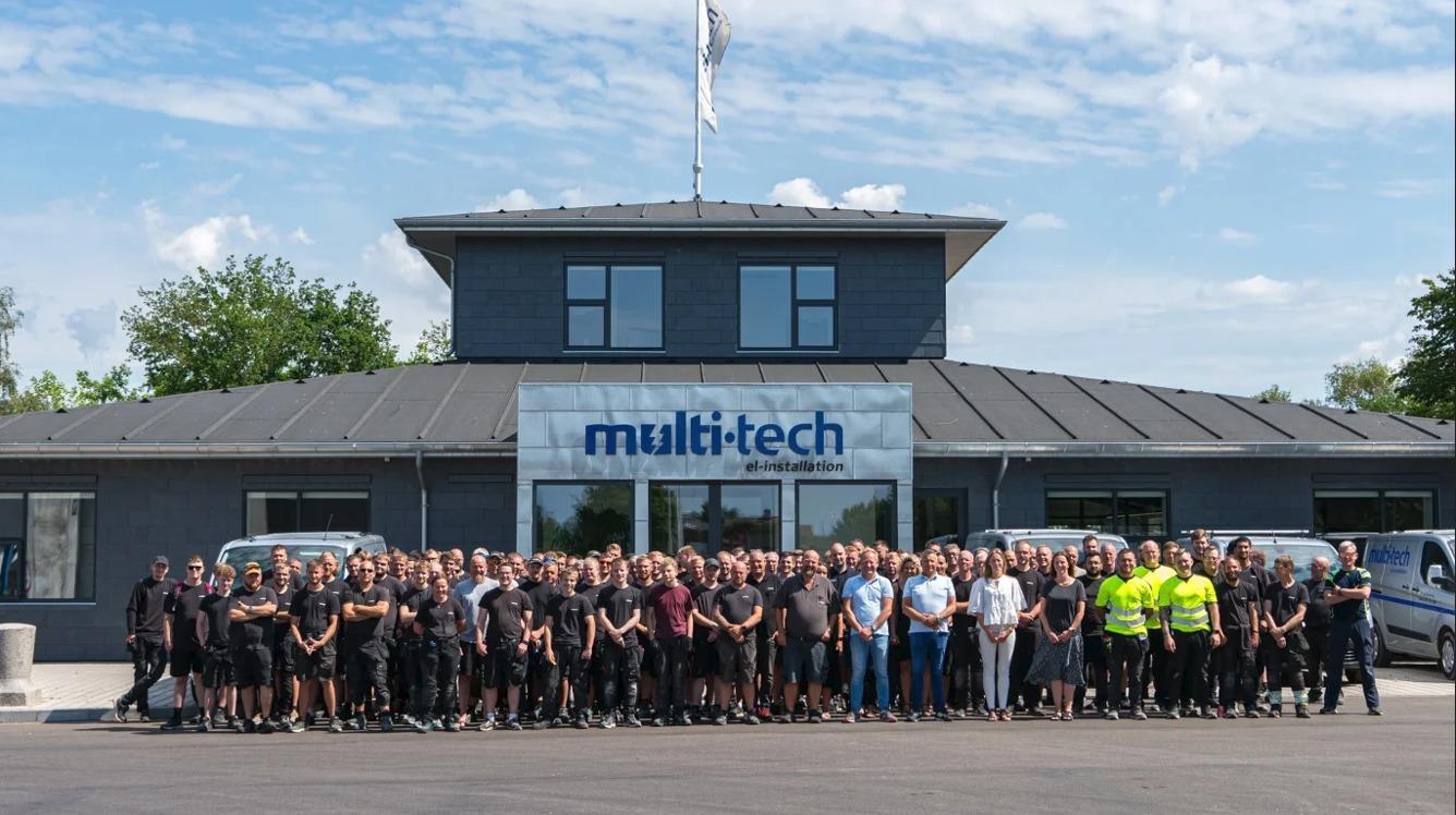 En stor gruppe medarbejdere står foran Multitech-bygningen på en solrig dag.
