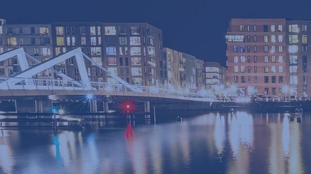 Aftenscene ved en bro over en kanal med oplyste bygninger og reflekser i vandet.