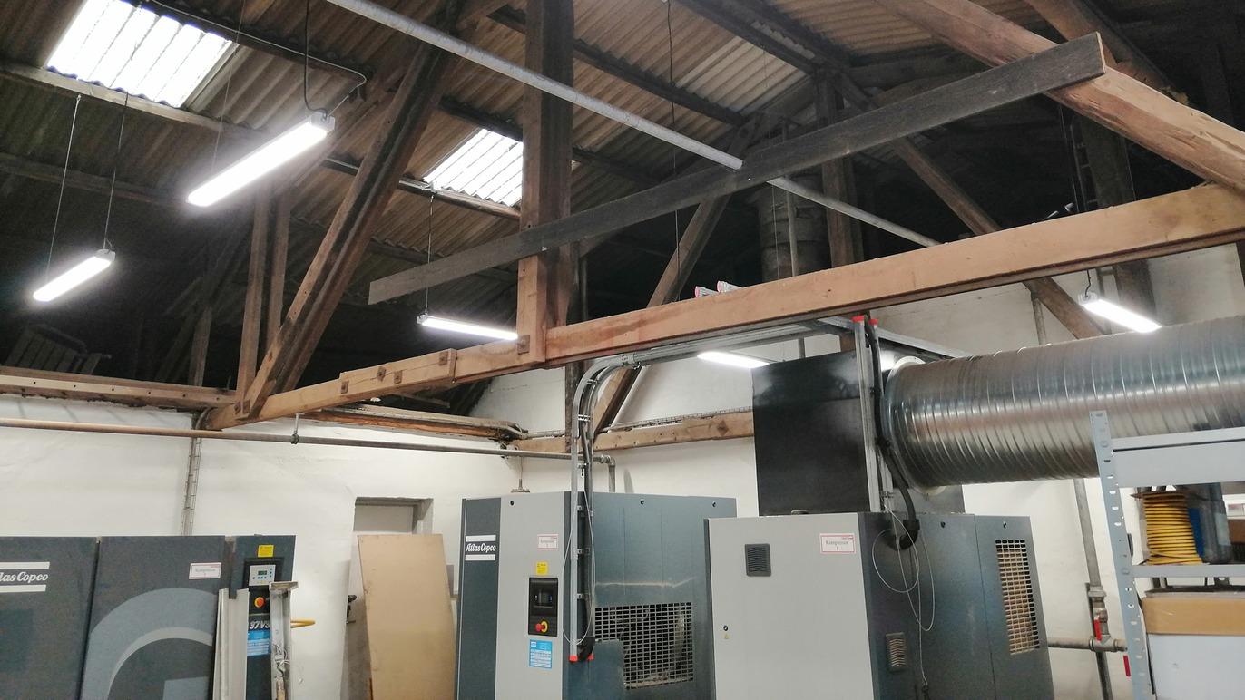 Industrielt værksted med maskiner, loftsbjælker og LED-belysning i loftet.