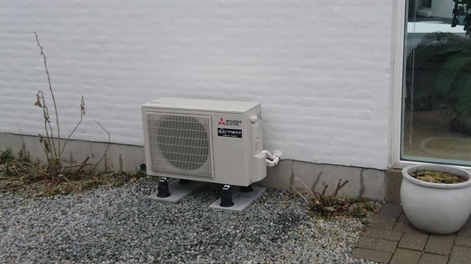 Udendørs klimaanlæg installeret ved en hvid væg, placeret på grå sten under et vindue.