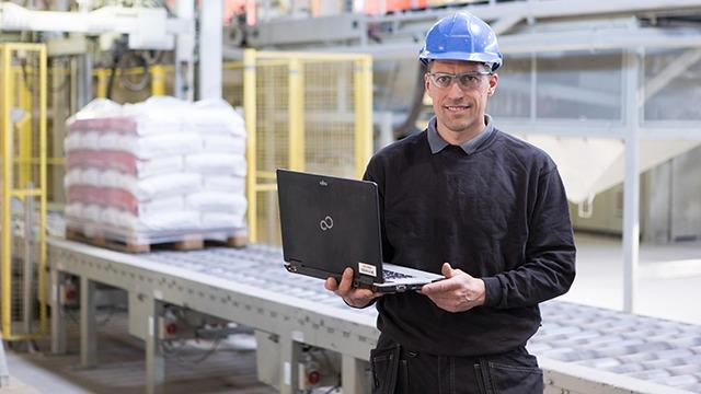 En mand i blå hjelm og beskyttelsesbriller står med en bærbar computer i en fabrik med en transportbånd i baggrunden.