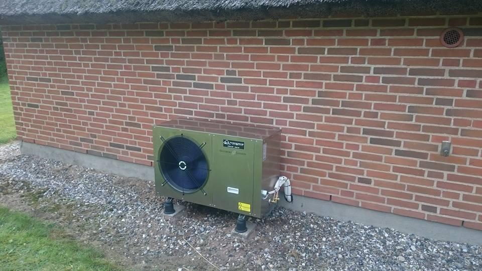 En varmepumpe installeret ved en murstensvæg med grus omkring, der viser en grøn enhed med en stor ventilator.