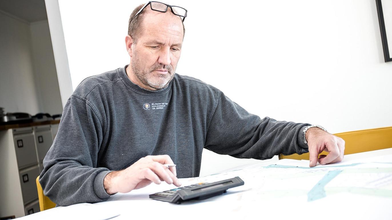 Mand sidder ved et bord med papirer og en lommeregner, mens han arbejder koncentreret.