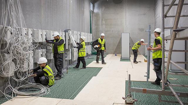 Arbejdsplads med elektrikere iført hjelme og sikkerhedsveste, der installerer kabler og udstyr i et rum.