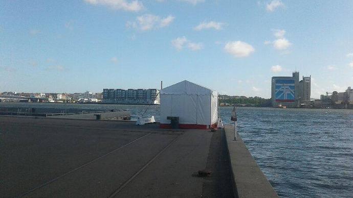 Hvid telt ved havnen med bygninger og vand i baggrunden under en blå himmel.