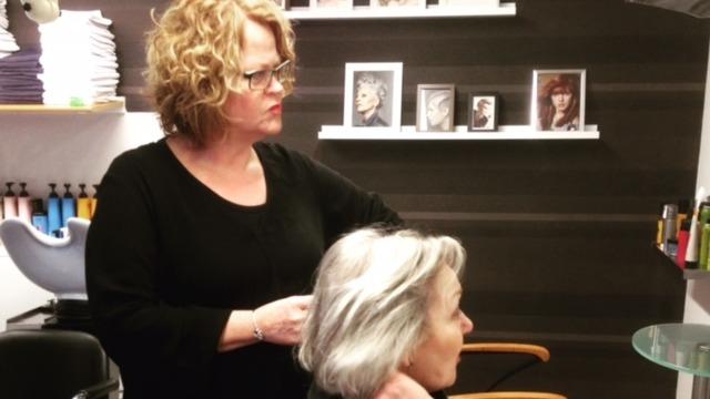 Frisør arbejder med en kvindes grå hår i en moderne salon med billeder på væggen.