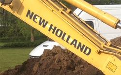 Gul gravemaskine fra New Holland, der arbejder på en byggeplads med jord og en hvid varevogn i baggrunden.
