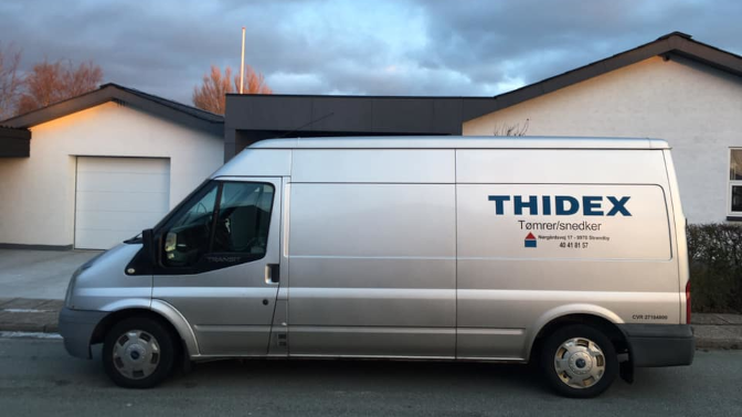 En sølvgrå varevogn med teksten "THIDEX Tømrerarbejde" parkeret foran et hus.
