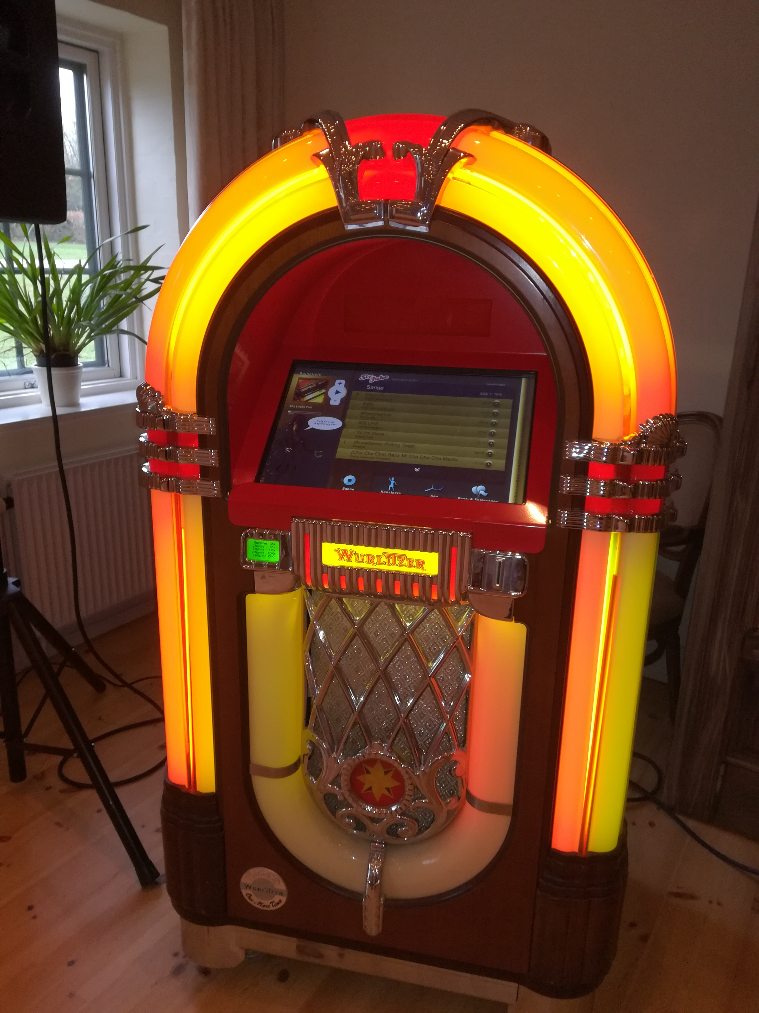 En farverig jukeboks med lysende detaljer og en berøringsskærm, placeret i et hyggeligt rum.