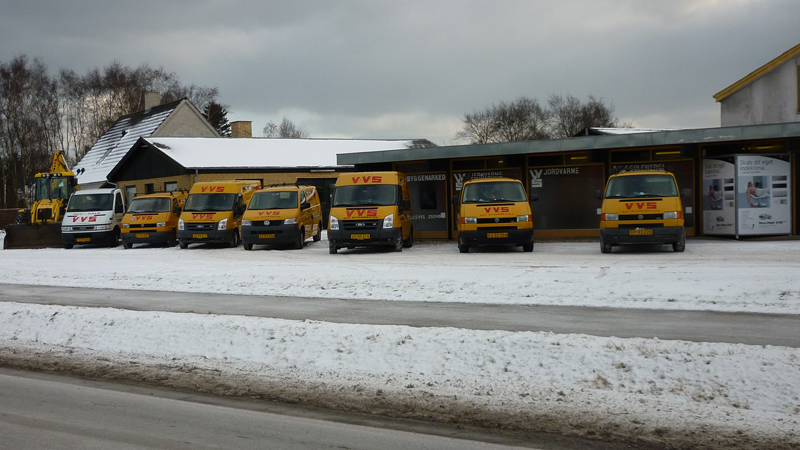 Syv gule varevogne parkeret foran en bygning i snevejr med en grå himmel.