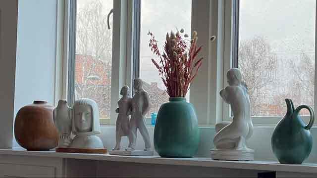 Vase med tørrede blomster og skulpturer på en vindueskarm, med forskellige keramiske genstande i baggrunden.
