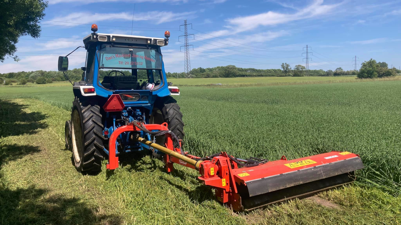 Traktor med en rød klipper arbejder på en grøn mark under en blå himmel med skyer.