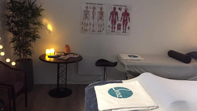 Behandlingsrum med massageborde, anatomiske plakater, stearinlys og en lille bord med magasiner.