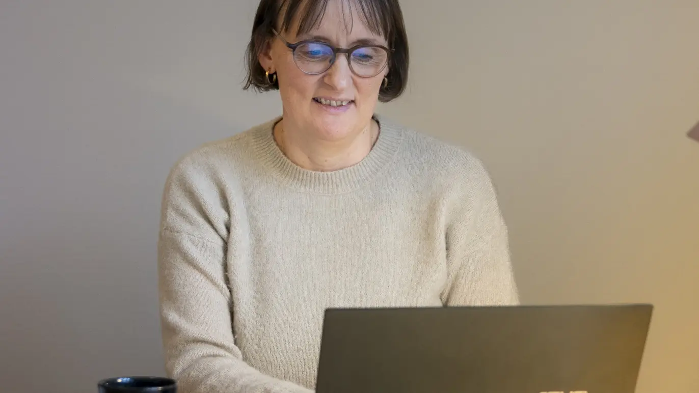 Kvinde i beige sweater sidder ved skrivebordet og arbejder på en bærbar computer med en kop kaffe ved siden af.