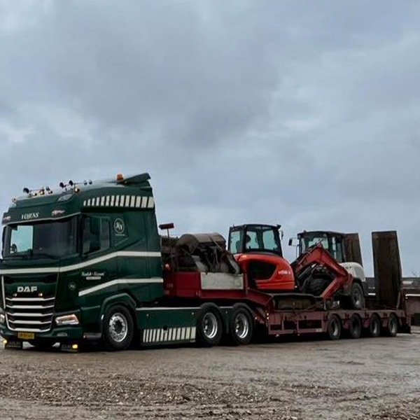 En DAF lastbil med en rød gravemaskine og en traktor på en trailer, parkeret på en grå dag.