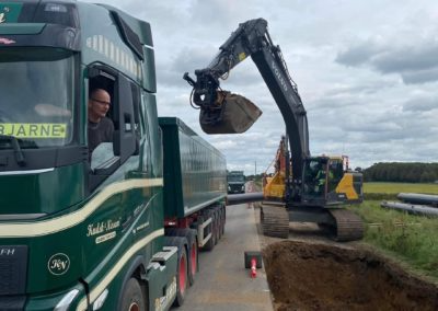 En lastbil med en chauffør og en gravemaskine, der fylder en container med jord ved en byggeplads.
