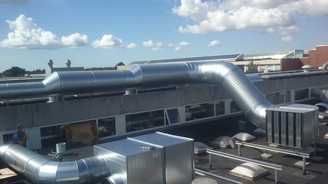 Billede af et tag med metalventilationskanaler og en stor ventilator på en bygning under en blå himmel.