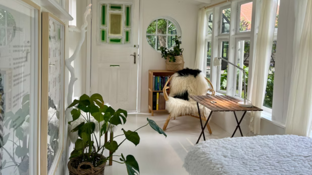 Lys og indbydende stue med planter, en stol, et bord og store vinduer, der giver udsigt til haven.