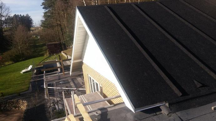 Taget på et hus med sort tagbelægning og hvidt gesims, omgivet af grønne træer og en have.