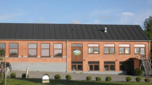En rød murstensbygning med sort tag og store vinduer, beliggende i et åbent område.
