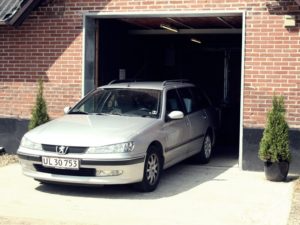 En sølvfarvet Peugeot stationcar parkeret foran en murstensgarage med små buske ved indgangen.