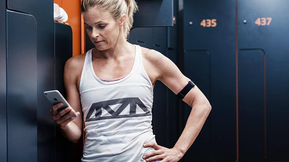 Kvinde i træningstøj står ved et skab og ser på sin smartphone i et fitnesscenter.