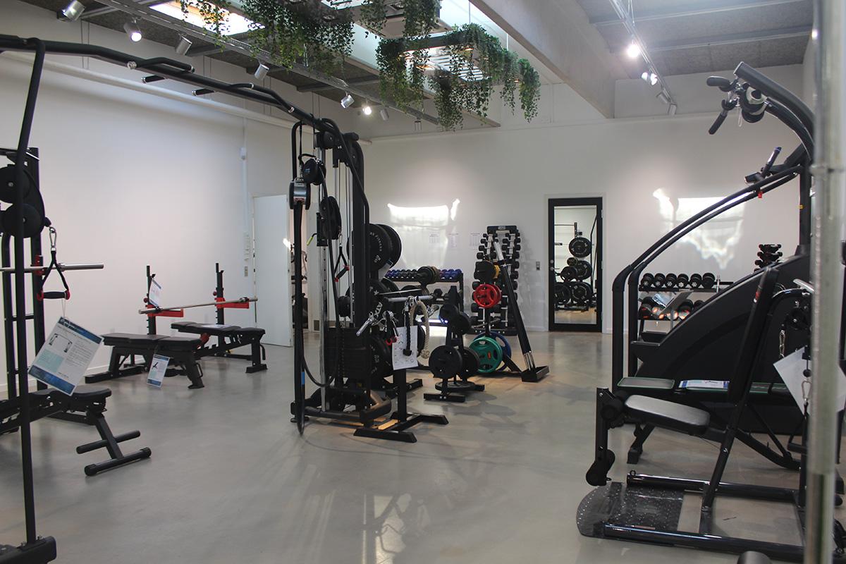 Træningslokale med forskellige fitnessmaskiner og vægte, herunder bænkpres og styrketræningsudstyr.
