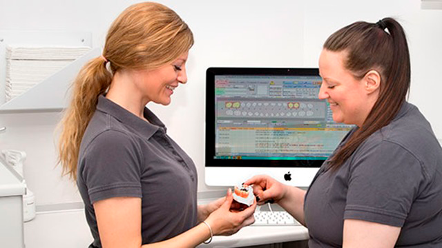 To kvinder i grå poloer, der smiler og diskuterer medicin ved en computer.