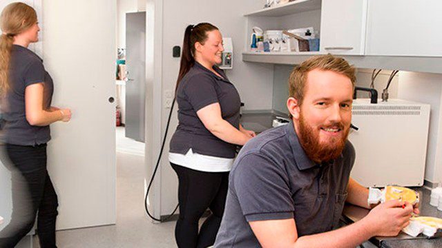 Tre personer i et moderne kontor, hvor en mand arbejder ved et skrivebord, mens to kvinder organiserer materialer.