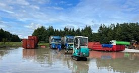 En lille gravemaskine arbejder i en vandfyldt byggeplads med lastbiler og containere i baggrunden.