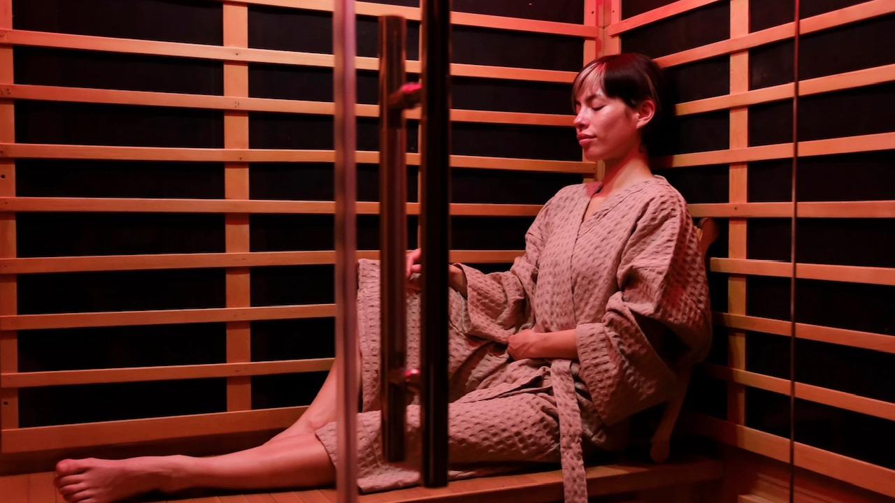Kvinde sidder afslappet i en sauna iført en badekåbe, omgivet af trævægge og blødt rødt lys.