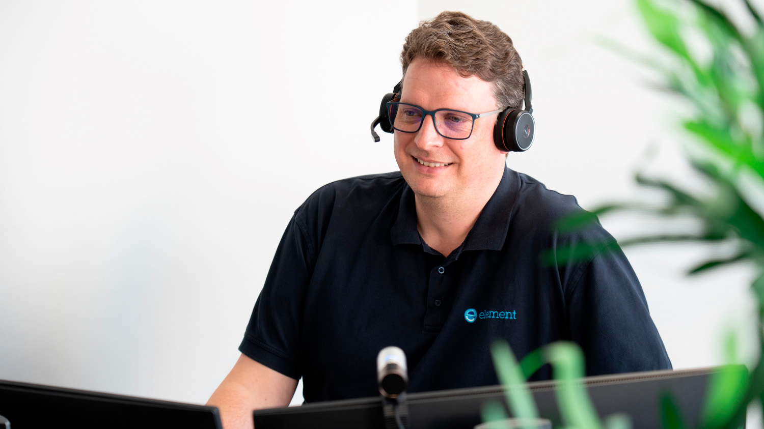 En mand med headset smiler, mens han arbejder ved en computer i et moderne kontormiljø.