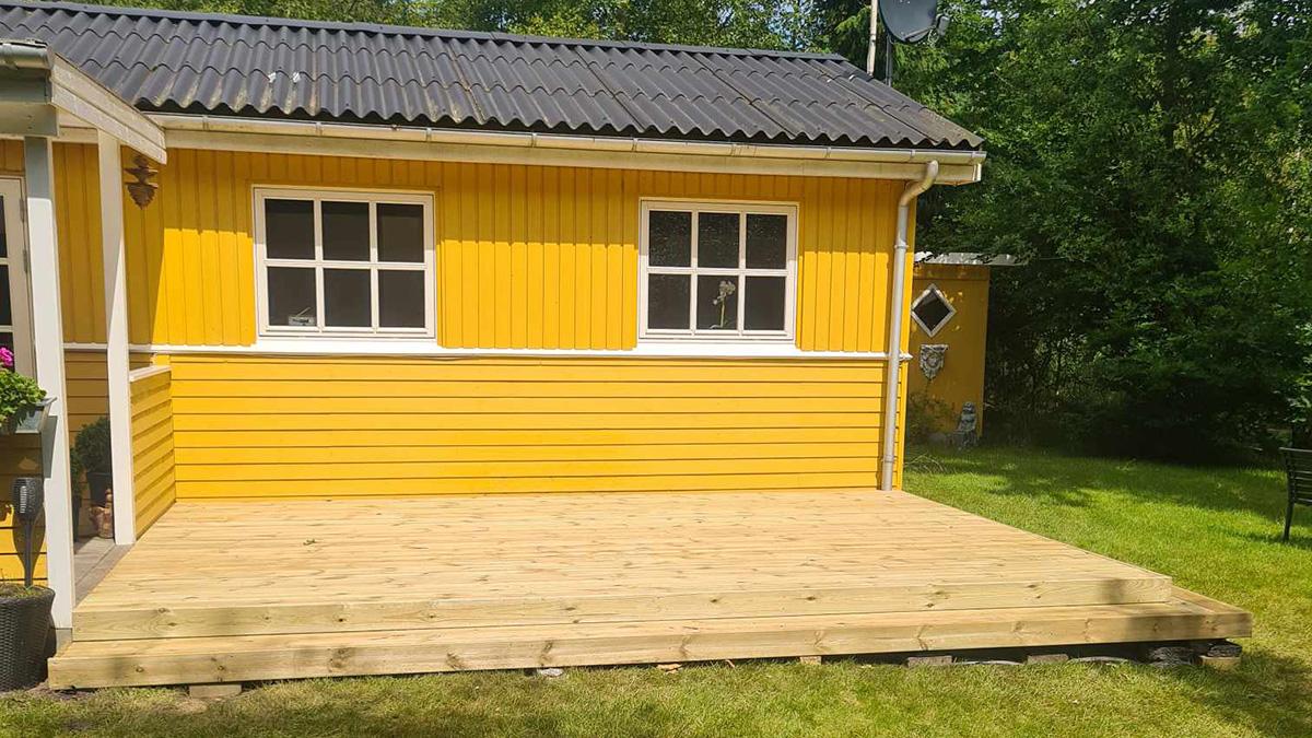 Gul træhytte med nye vinduer og en træterrasse i haven, omgivet af grønne træer.