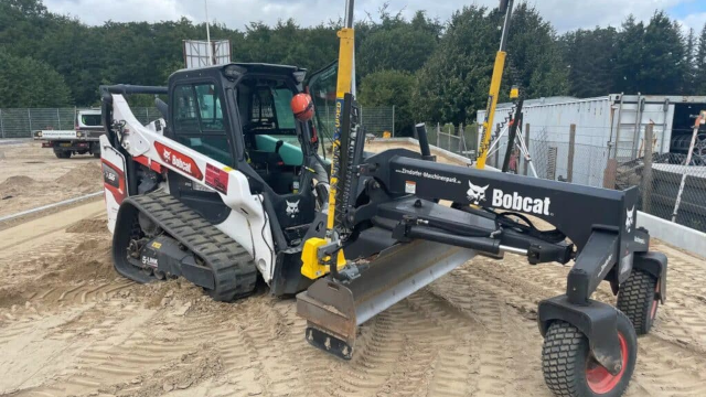 En Bobcat minilæsser med skovl står på en byggeplads omgivet af sand og træer.
