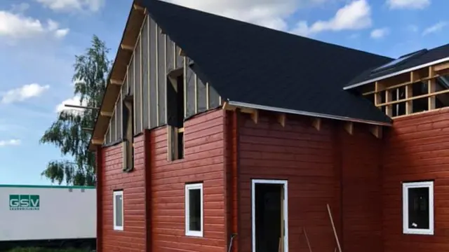 Nybygget hus med rød træbeklædning og sort tag under en blå himmel. Byggeplads med materialer i baggrunden.