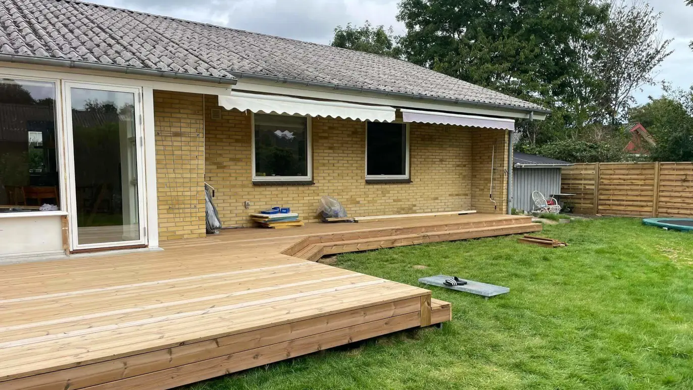 Husets baggård med træterrasse, græsplæne og vinduer, der giver udsigt til haven.