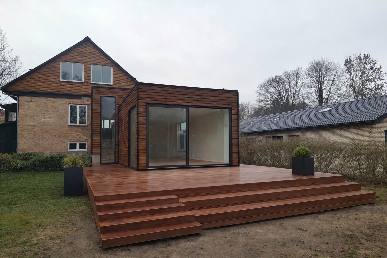 Moderne træhus med store vinduer og terrasse i haven, omgivet af grønne planter og træer.