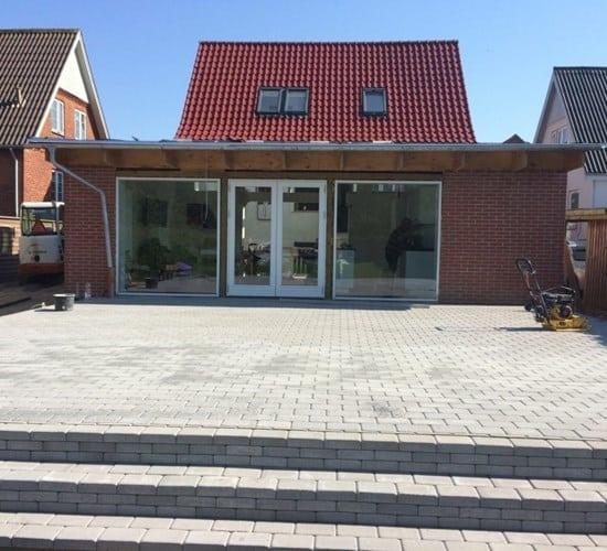 Moderne hus med rød tag og store vinduer, beliggende på en flisebelagt terrasse i solrigt vejr.