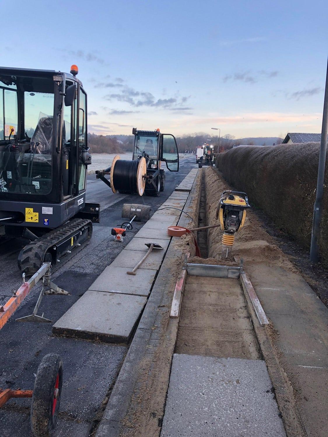 Biler og maskiner arbejder på en vej, hvor der lægges fliser og graves en grøft til dræning.