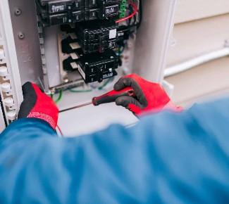 En person i blå jakke arbejder med en elektrisk tavle og bruger en skruetrækker iført røde handsker.