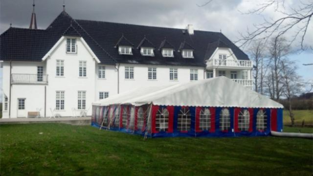 Hvidt hus med sort tag og festtelt i haven, omgivet af grøn græsplæne og træer.