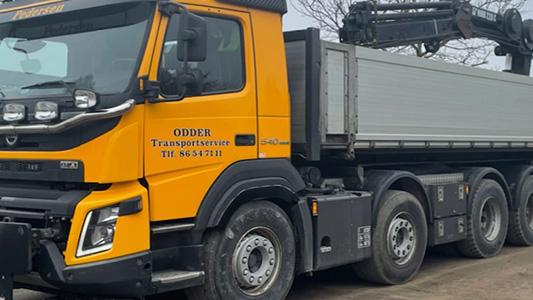 En stor gul lastbil fra Odders Transport med en grå lad og kran, parkeret på en grusvej.