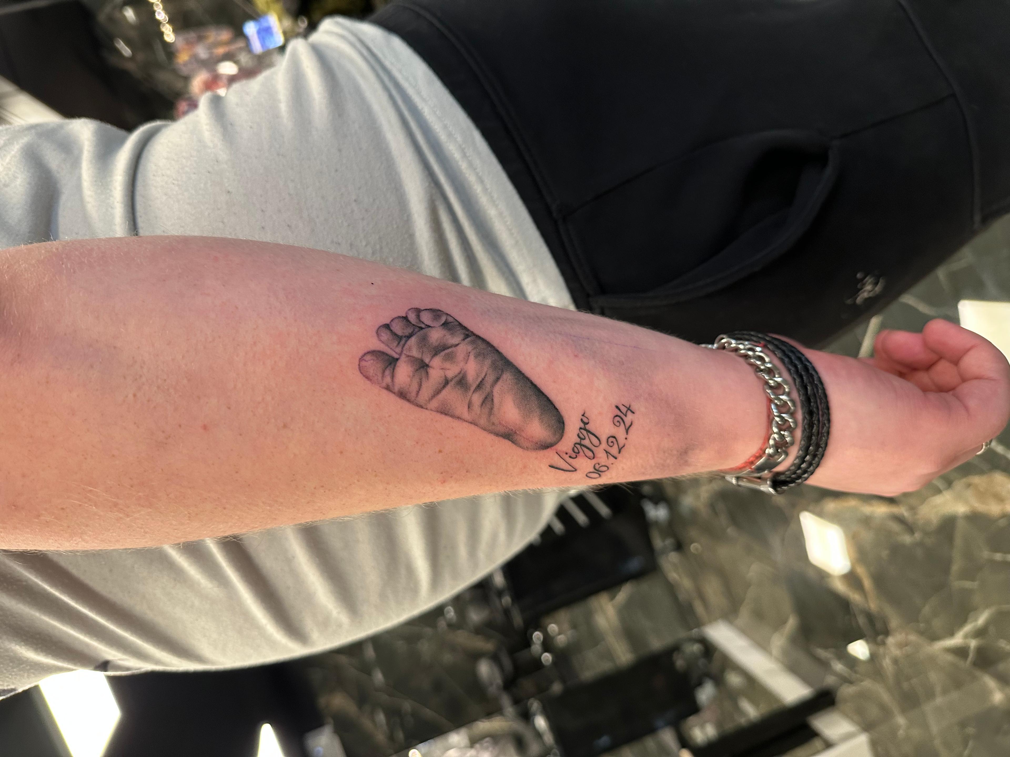Tatovering af en babys fodaftryk på en mands underarm med teksten "Forever in my heart".