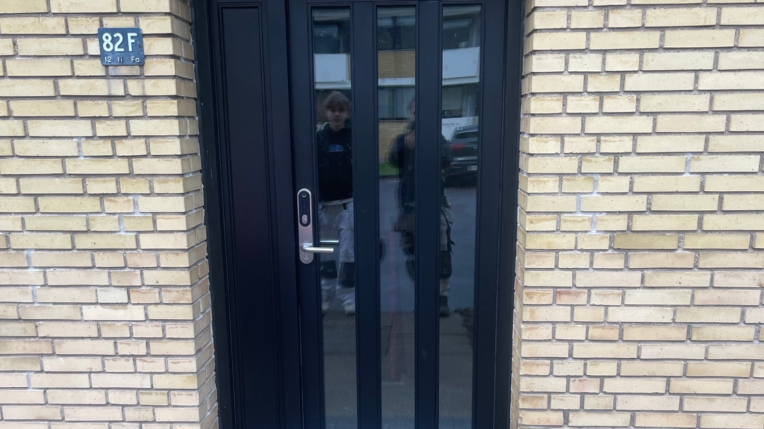 En sort dør med glaspartier og en person, der står foran, i en murstensbygning.