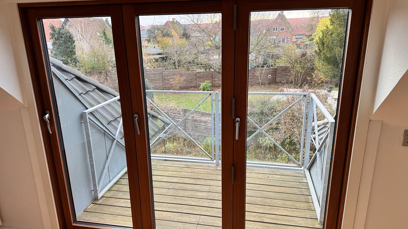 Udsigt fra en balkon med trædæk og metalgelænder, omgivet af grønne træer og bygninger.