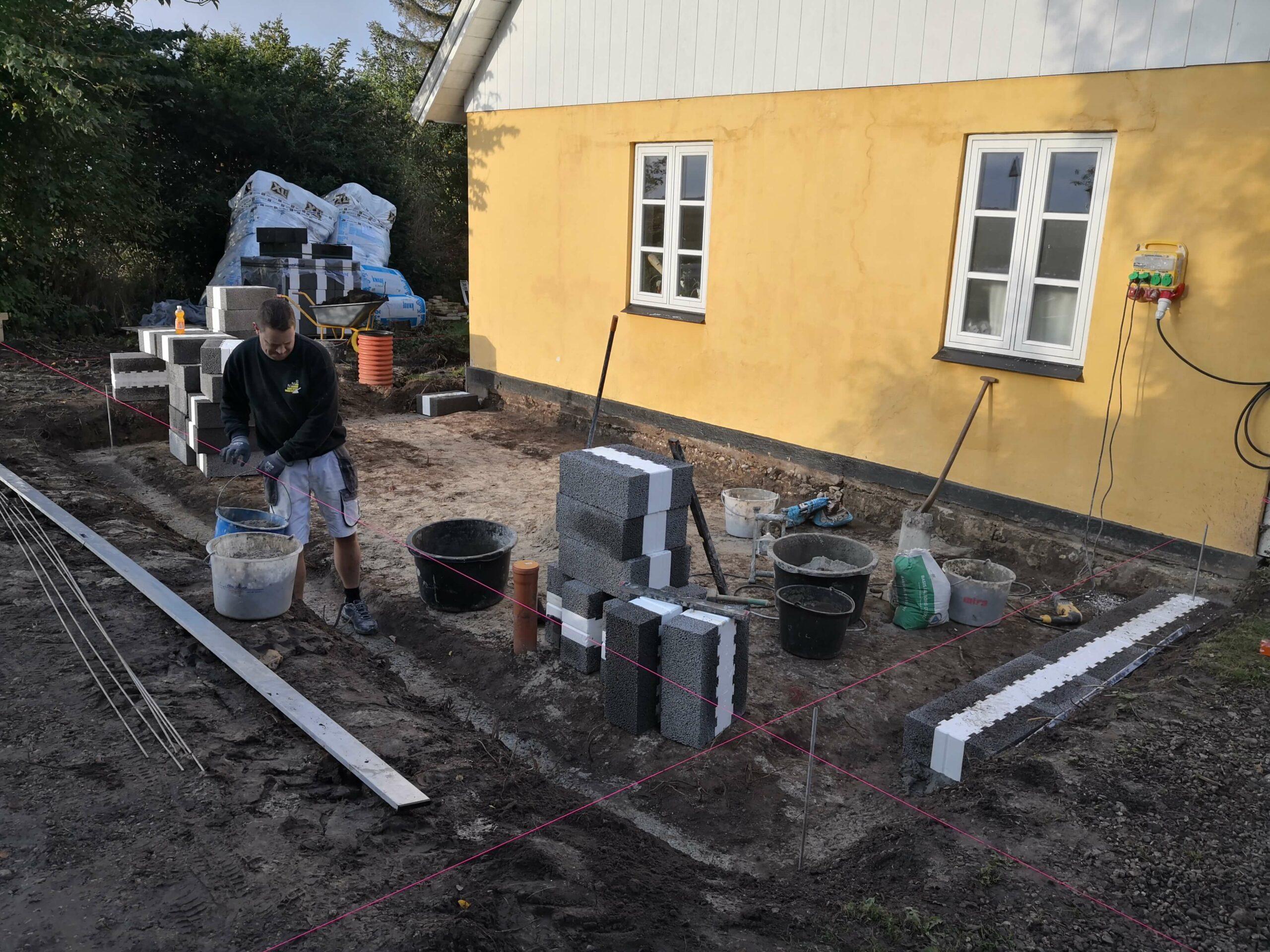 En håndværker arbejder på en byggeplads ved en gul husvæg med mursten og værktøj spredt omkring.