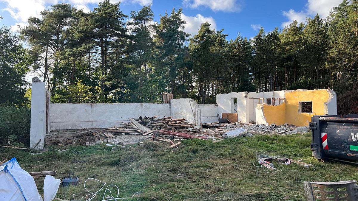 Nedrevet hus med synlige vægge og affald i en skovklædt område under en blå himmel.
